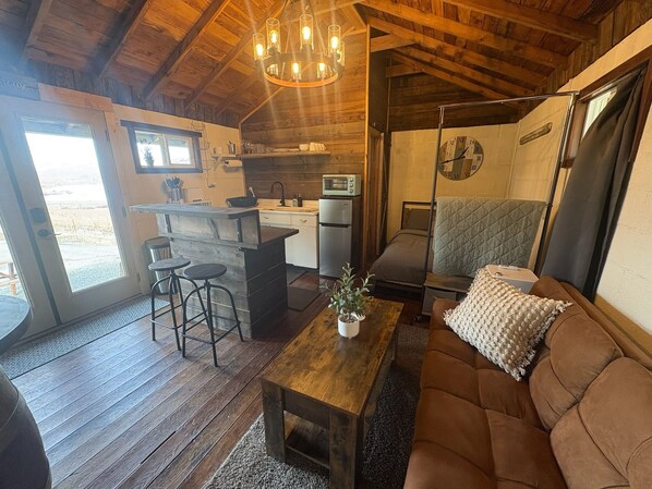 L’intérieur chaleureux d’un chalet avec un plafond en bois, un coin cuisine, un espace salon avec un canapé et des tabourets de bar, et une grande horloge au mur.