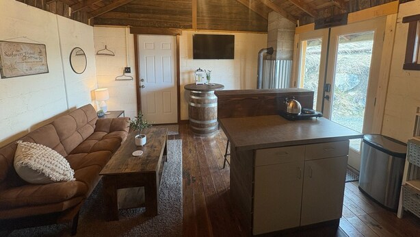 A cozy cabin interior with a wooden ceiling, a sofa, a coffee table, a kitchen area with a barrel, and a door leading outside.