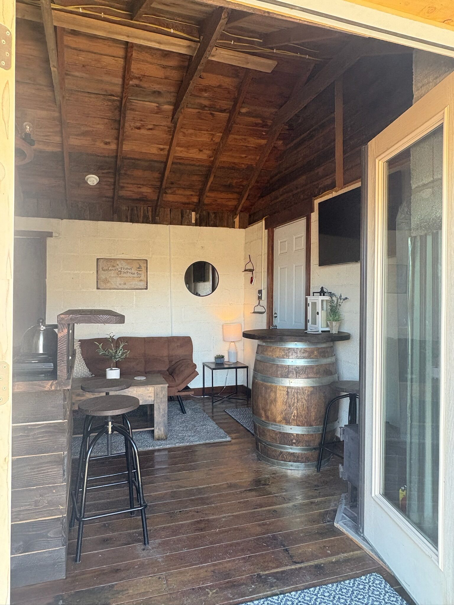A rustic interior with a wooden ceiling, a round mirror, a sofa, a coffee table, a bar stool, and a large wooden barrel.