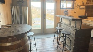 A rustic kitchen with a wooden bar, stools, and a view of a mountainous landscape through the door.