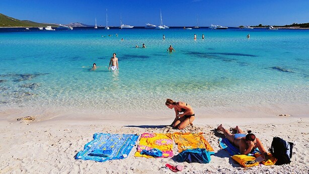Una playa con agua turquesa cristalina, gente nadando y tomando el sol, y barcos a lo lejos.