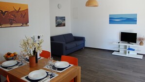 Un comedor con una mesa de madera dispuesta para dos personas, un sofá azul y un mueble bajo para televisor con una televisión de pantalla plana.