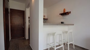 Ein Korridor mit einer Holztür, eine Küchenzeile mit einem Regal, auf dem eine Uhr und Gläser stehen, sowie zwei weiße Barhocker.