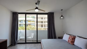 Ein Schlafzimmer mit Bett, Schreibtisch, Stuhl, einem Balkon mit Blick auf Gebäude und einem Deckenventilator.