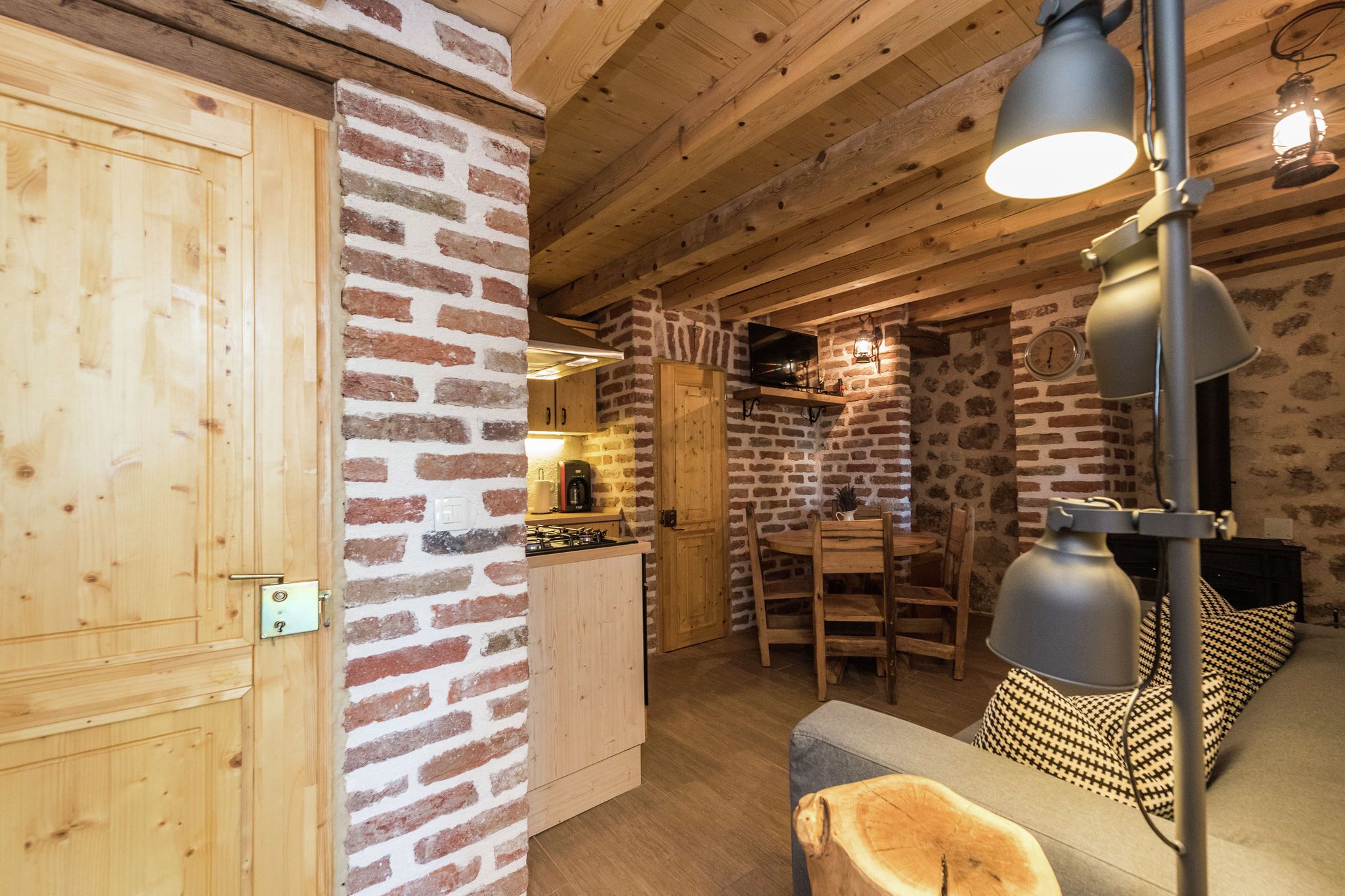 A rustic interior with exposed wooden beams, a brick wall, a wooden door, a dining area with a table and chairs, and a sofa with patterned cushions.