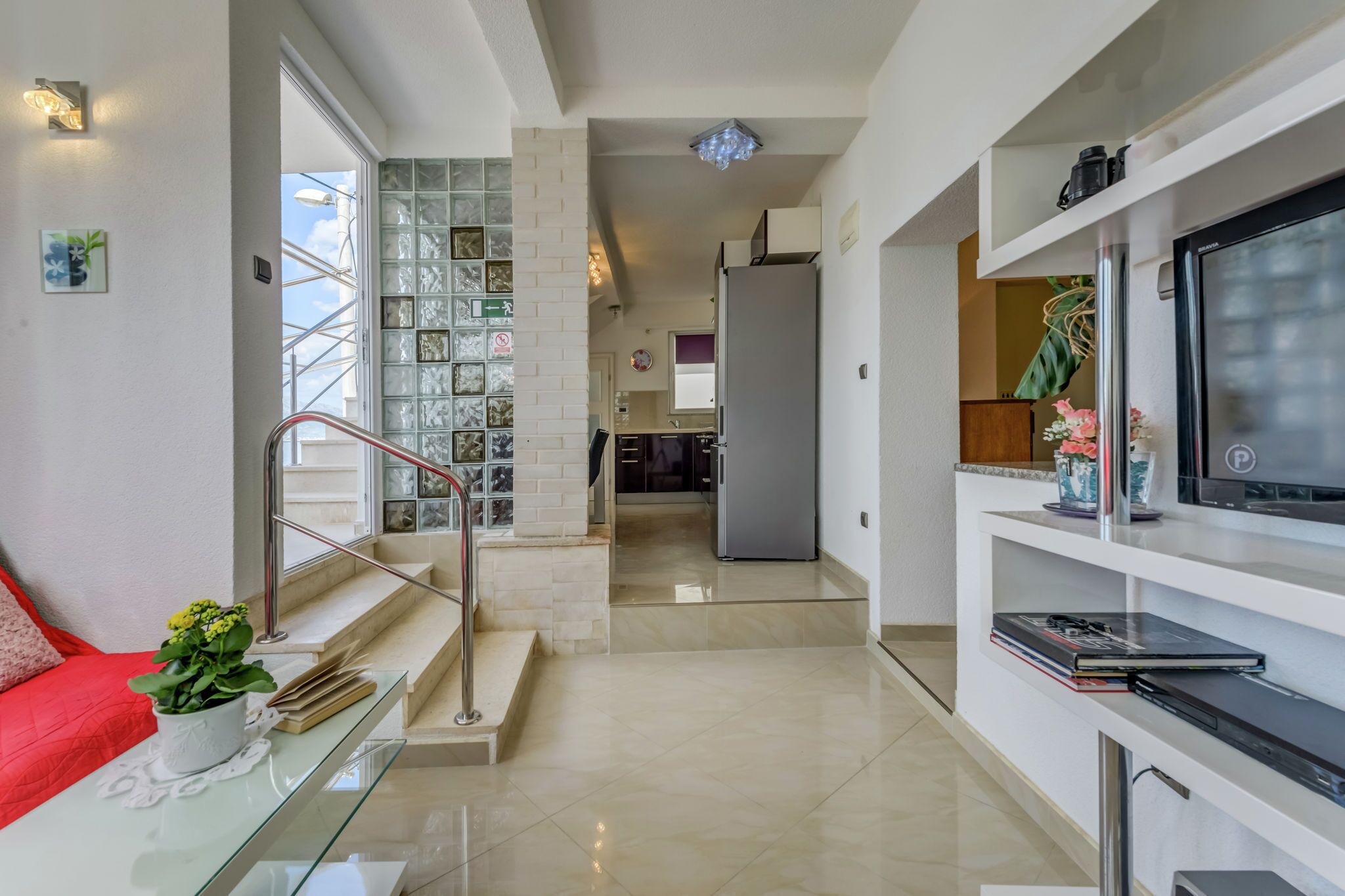 A modern interior with a glass staircase, a flat-screen TV, and a kitchen area.