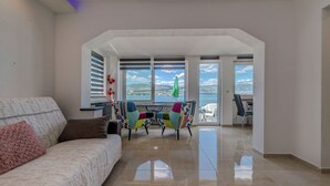 Un salon moderne avec un canapé, des fauteuils colorés, une table à manger et une vue sur la mer grâce à de grandes fenêtres.