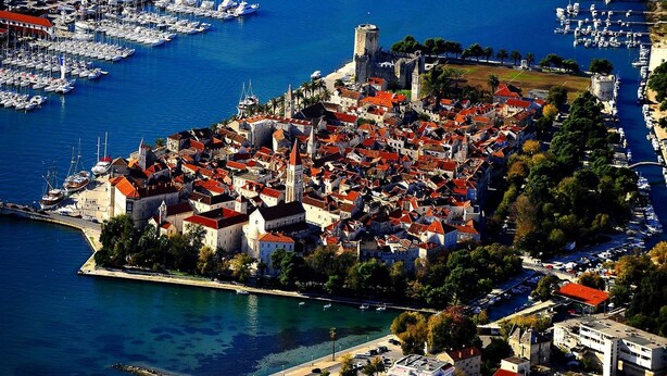Vue aérienne d’une ville côtière avec un château historique, un port de plaisance et une zone urbaine dense, caractérisée par des bâtiments aux toits rouges.