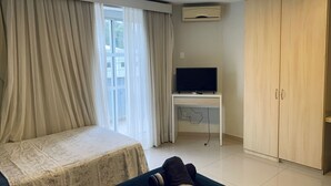 A bedroom with a bed, a TV on a stand, a wardrobe, and a window with curtains. - Saint Sebastian ap103-stúdio  (Jaragua do Sul)