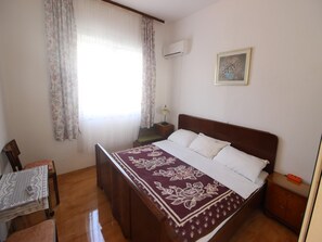 Une chambre à coucher avec un lit, une table de chevet, une lampe, une chaise, une table recouverte d’une nappe en dentelle et une fenêtre avec des rideaux.