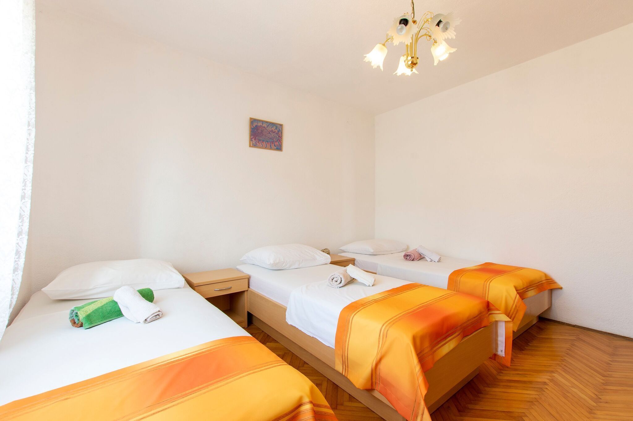 A hotel room with three single beds, each with white bedding and orange blankets. There is a small painting on the wall and a chandelier above the beds.