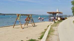 A beach with a swing set, a thatched-roof structure, and a paved walkway along the water. - Apartment in Ljubac with Barbecue (Ljubac)