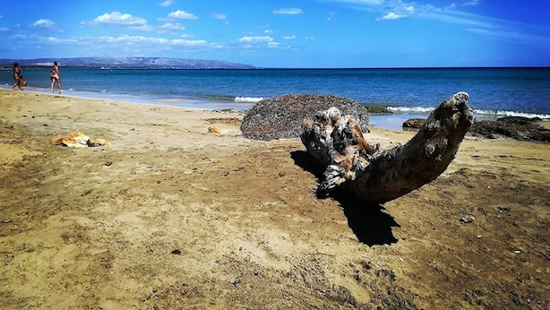 Une plage avec une grande formation rocheuse au premier plan, un ciel bleu dégagé et quelques personnes au loin.