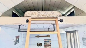 Ein Schlafpodest mit einer Holzleiter, ein Kissen mit Blumenmuster und ein Schild mit motivierenden Sprüchen an der Wand.