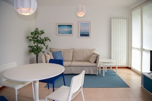 A modern living room with a round white table, a beige sofa, and a blue rug. There are two framed pictures on the wall, a potted plant, and a radiator.
