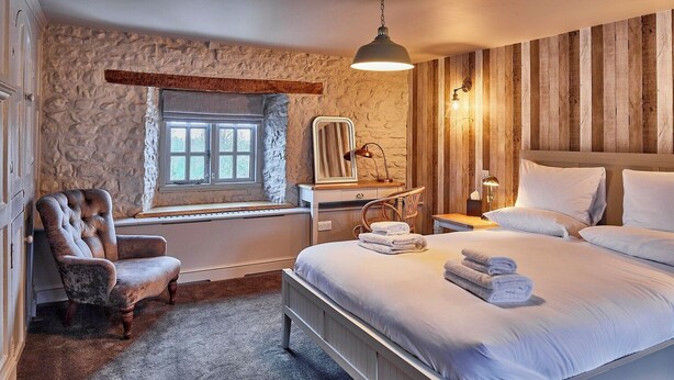 Une chambre avec un mur en pierre, un grand lit, une fenêtre, une chaise et une tête de lit en bois.