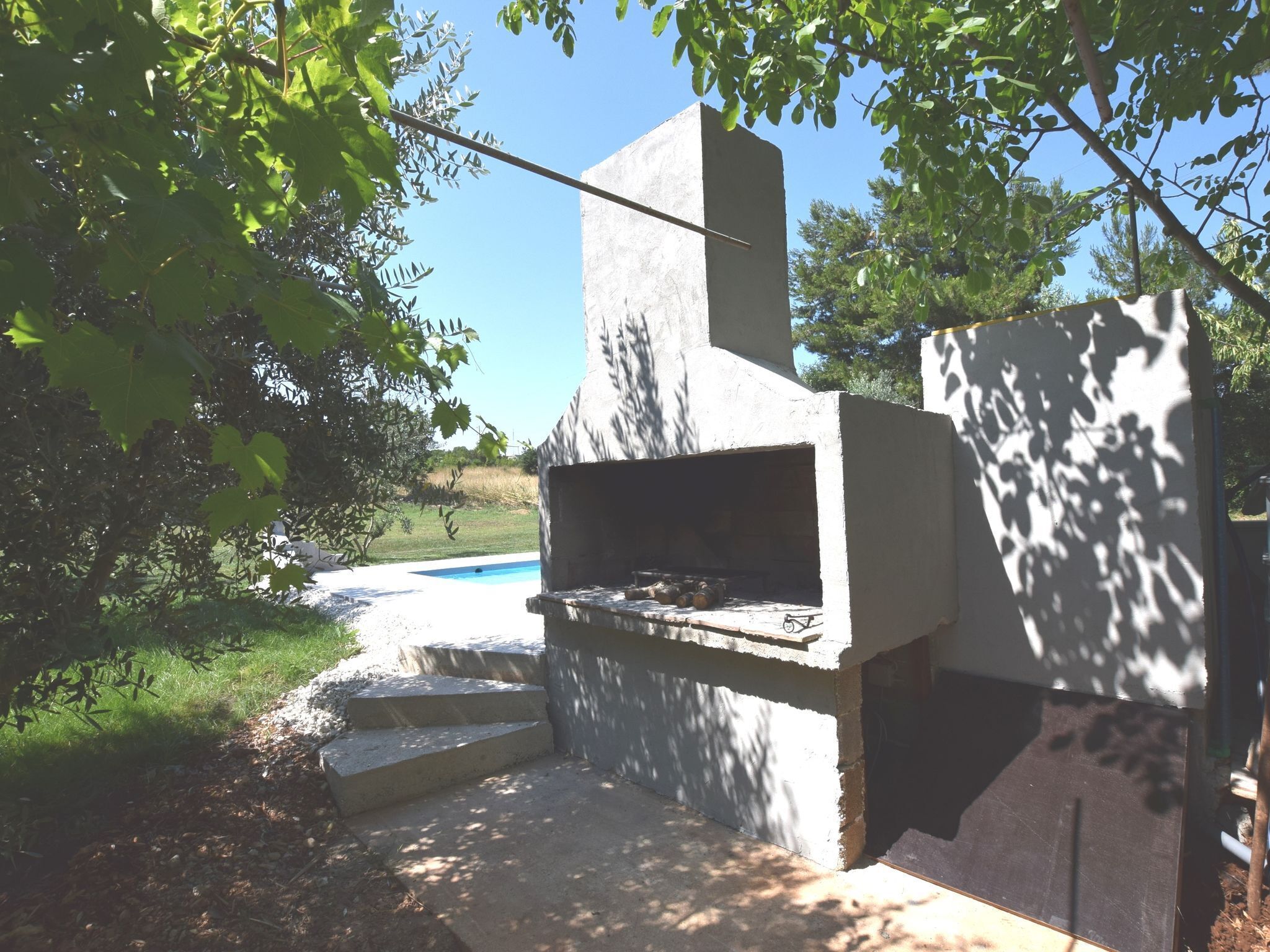 Uma churrasqueira ao ar livre com estrutura de concreto, degraus que a levam e uma piscina ao fundo.