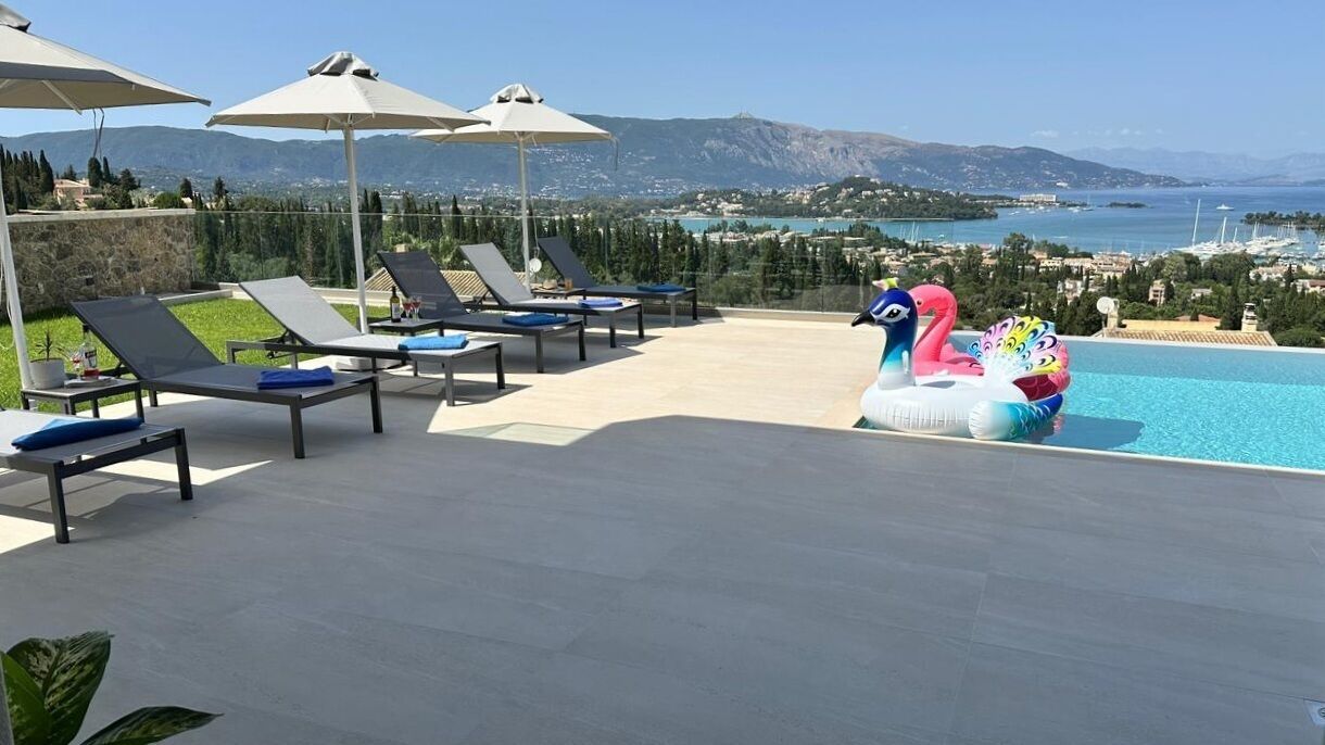 A poolside area with lounge chairs and umbrellas, a decorative flamingo float, and a scenic view of mountains and a body of water.