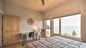 A bedroom with a large bed, a wooden door, a round mirror, a desk with a chair, and a view of the sea through large windows.