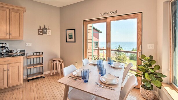 A dining area with a table set for four, a view of the sea through large windows, and a kitchen area with wooden cabinets and a cart.