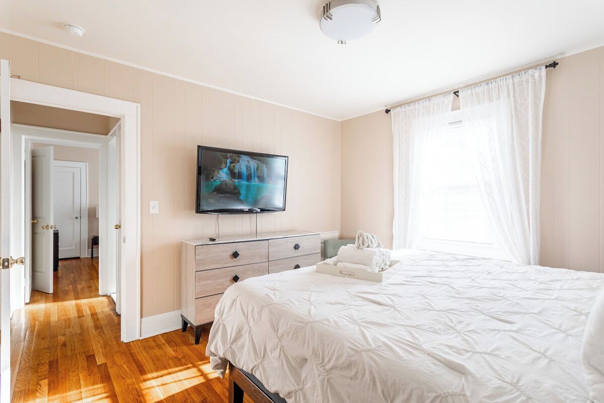 Un dormitorio con cama, televisor montado en la pared, cómoda y ventana con cortinas.