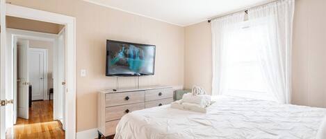 Un dormitorio con cama, televisor montado en la pared, cómoda y ventana con cortinas.