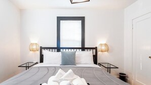 A neatly made bed with rolled towels, two bedside tables with lamps, a window with blinds, and a door. - Wells Whole House (Bridgeport)