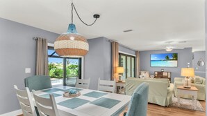 Un salon moderne avec un coin repas, doté d’une grande fenêtre offrant une vue sur la verdure, d’un luminaire suspendu et d’un ventilateur de plafond.