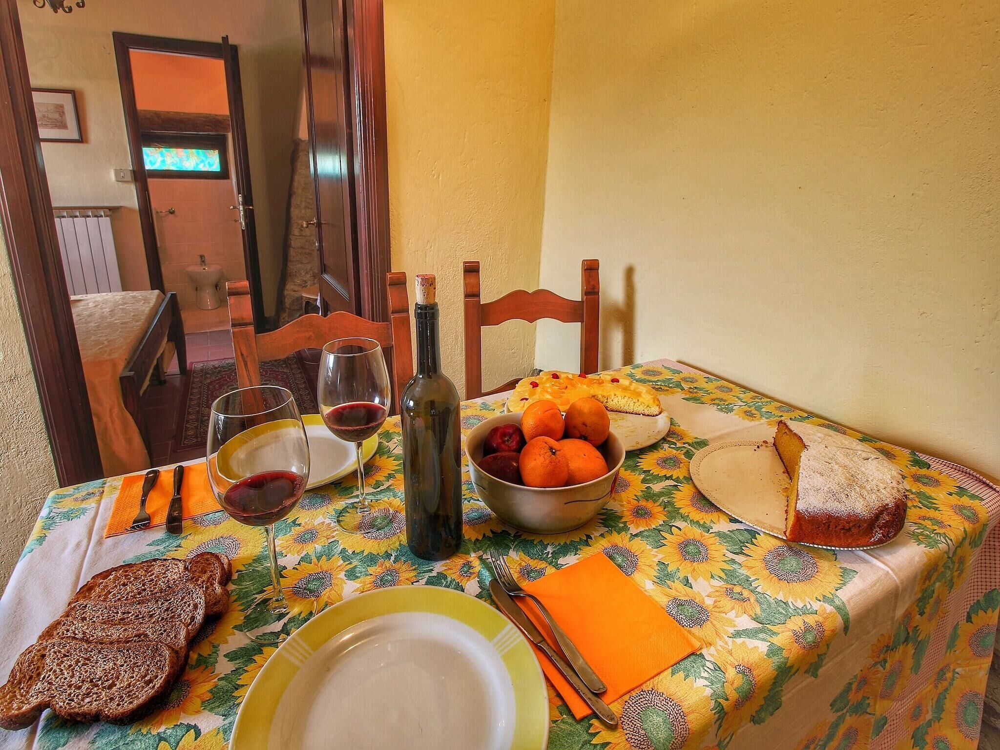 A dining table with a floral tablecloth, a bottle of wine, a bowl of fruit, and slices of bread.