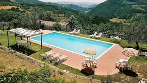 Ein rechteckiger Swimmingpool mit Liegestühlen und Sonnenschirmen, umgeben von üppiger Vegetation und Hügeln.