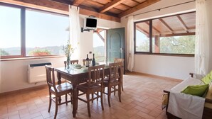 A dining area with a wooden table and chairs, a TV mounted on the wall, and large windows offering a view of the outdoors. - Farmhouse in Apecchio with Panoramic Views (Apecchio)