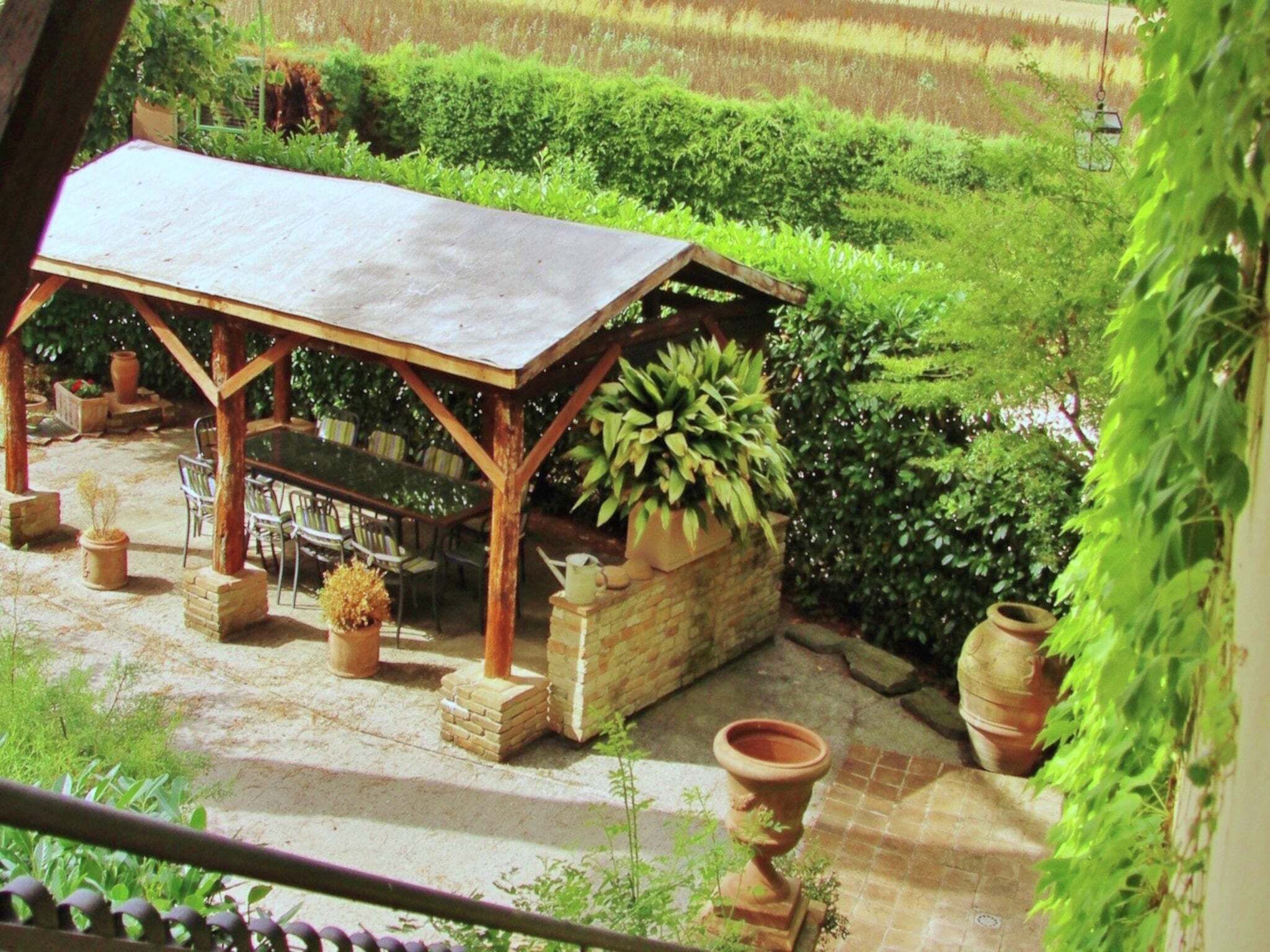 A covered outdoor seating area with a table and chairs, surrounded by potted plants and a brick planter.