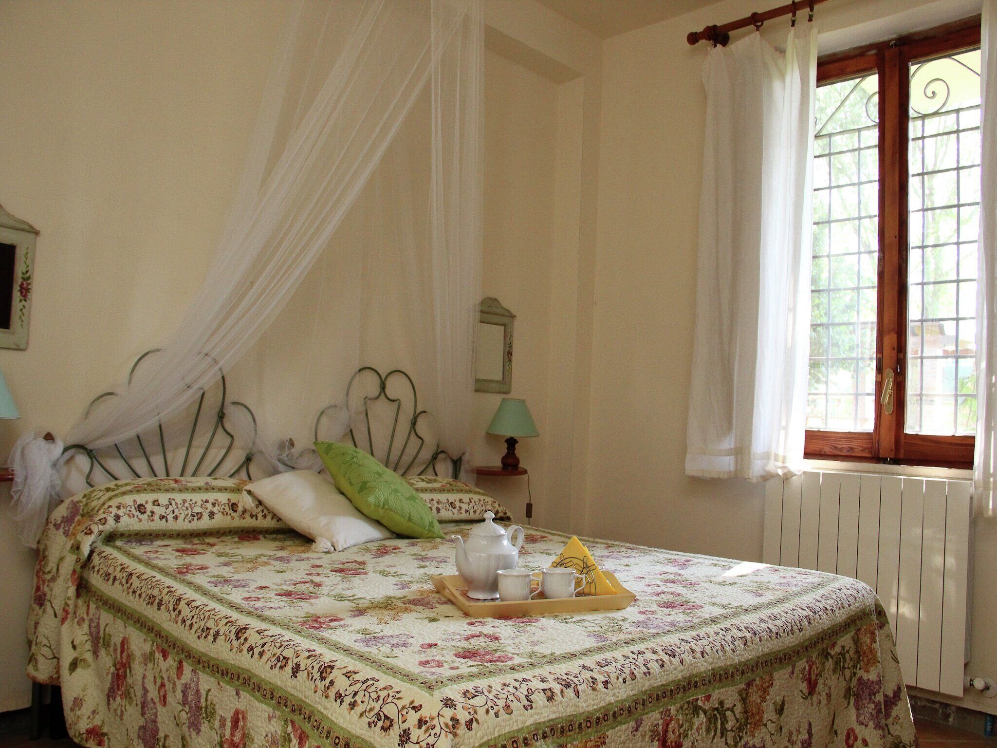 Ein Bett mit floralem Überwurf, ein Nachttisch mit Lampe und ein Fenster mit Vorhängen.