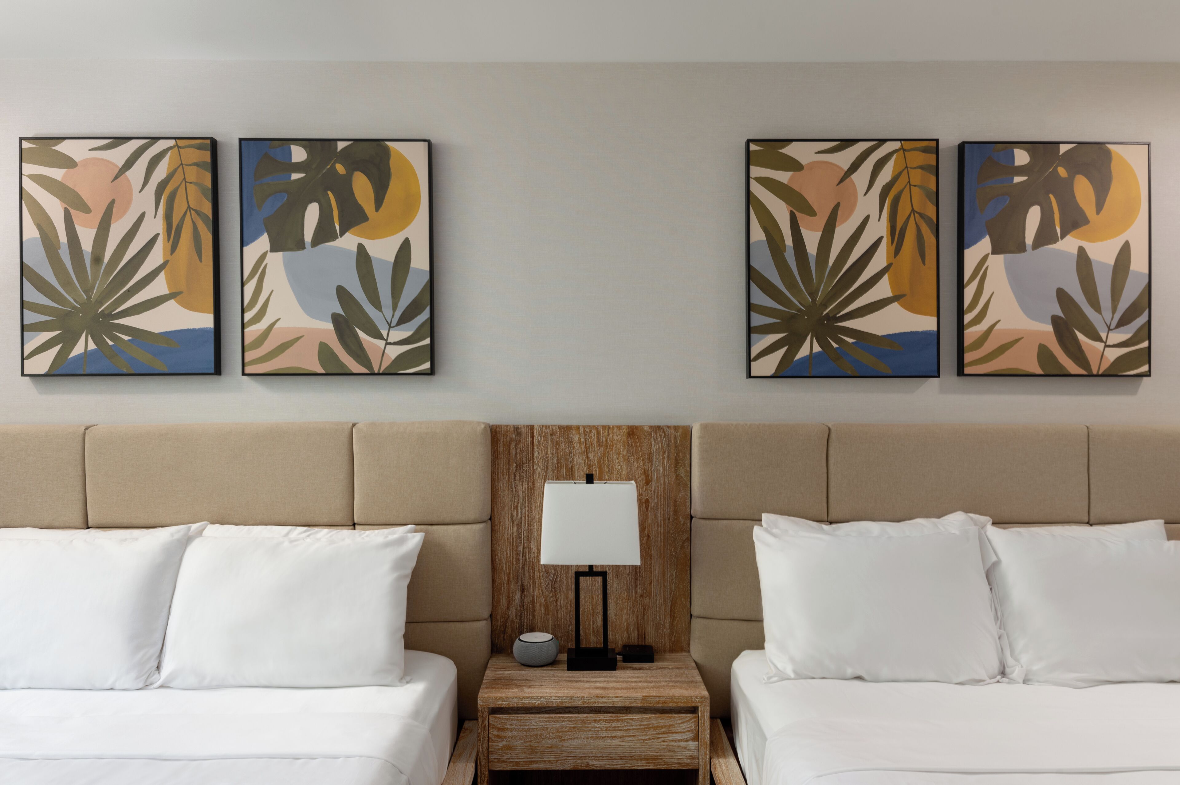 A hotel room with a wooden headboard, two beds with white linens, a nightstand with a lamp, and four abstract paintings on the wall.