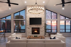 A spacious living room with a stone fireplace, large windows, and a ceiling fan. - The Wave at Marblehead (Marblehead)