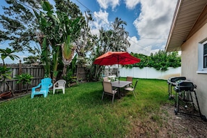 A backyard with a dining table, chairs, a red umbrella, and a barbecue grill.