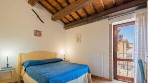 A bedroom with a wooden ceiling, a bed with a blue and beige bedspread, a nightstand with a lamp, and a window with a view of a building outside. - Holiday Home in Sellano with Pool & Sun Terrace (Sellano)