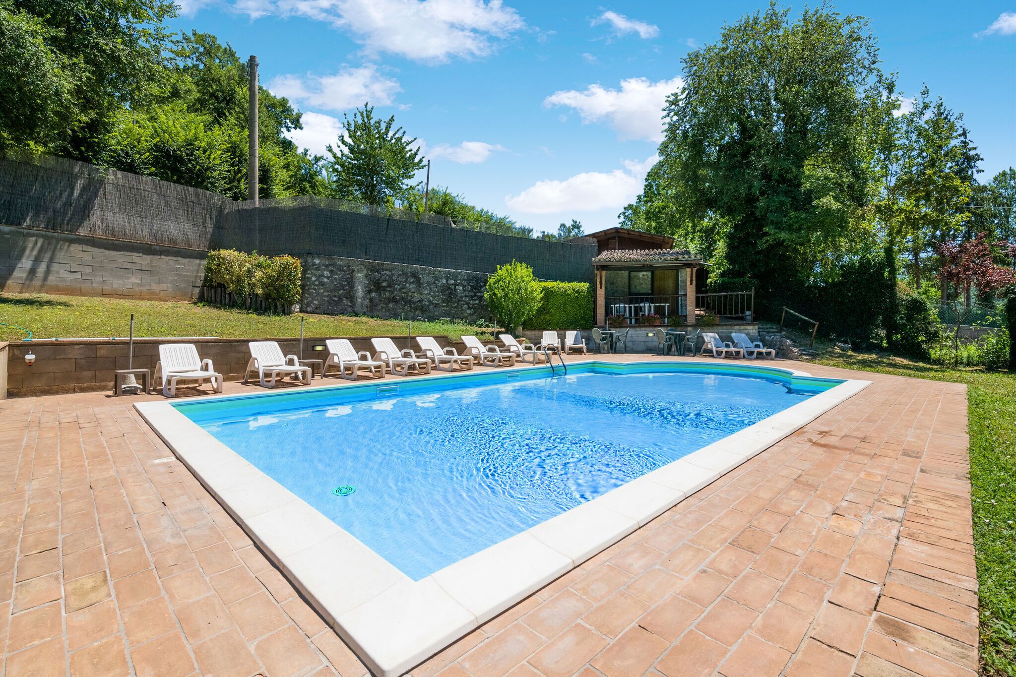 Ein rechteckiger Pool mit gefliestem Sonnendeck, umgeben von weißen Liegestühlen und einer überdachten Sitzgruppe.