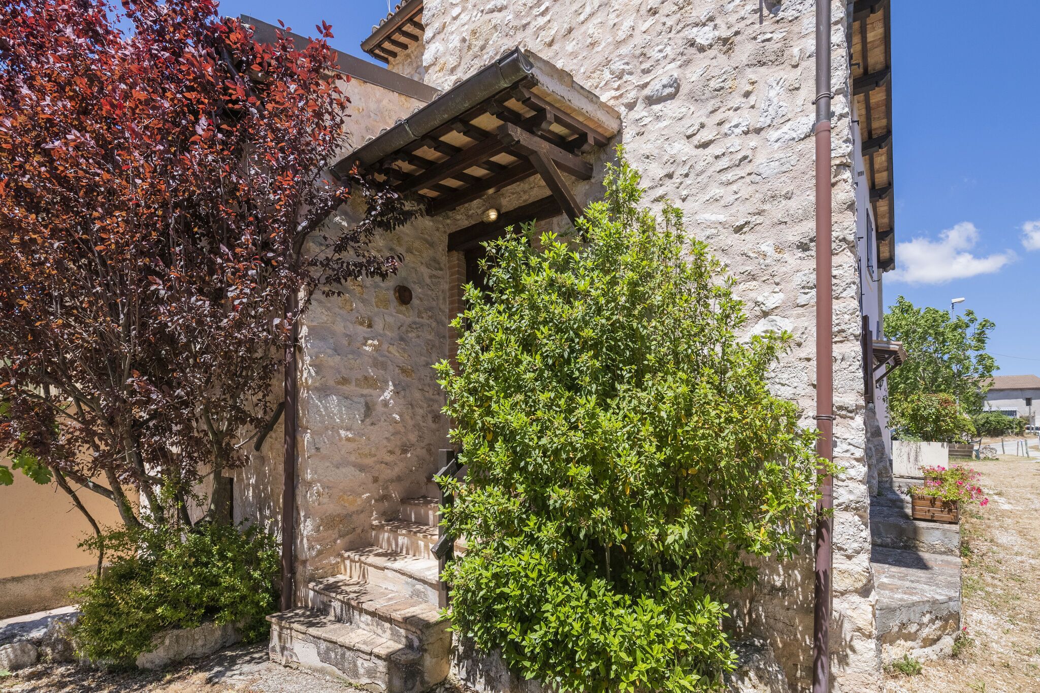 A stone building with a wooden staircase leading to a door, surrounded by greenery and a clear sky.