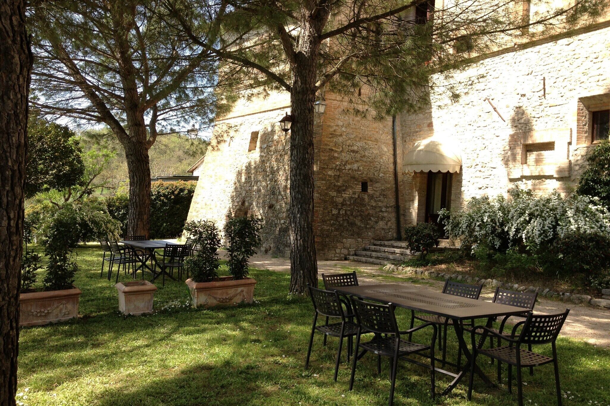 An outdoor dining area with a stone building in the background, surrounded by trees and potted plants.