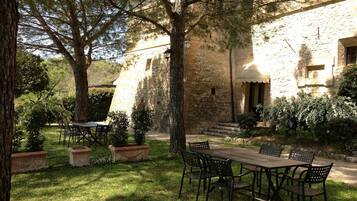 An outdoor dining area with a stone building in the background, surrounded by trees and potted plants.