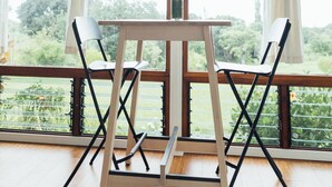 Una habitación con una mesa de madera y dos sillas, un florero con flores y ventanas grandes con cortinas.
