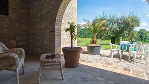 Une terrasse extérieure avec des murs en pierre, une chaise en osier, une petite table recouverte d’une nappe et des plantes en pot.