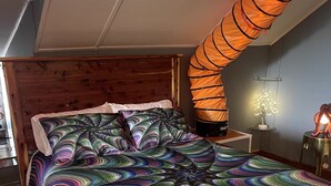 Un dormitorio con cabecera de madera, edredón colorido a rayas, una original lámpara de pantalla naranja y una mesita de noche con una lámpara pequeña.