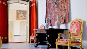 Una habitación con una cortina roja grande, un tapiz estampado, una mesa de madera oscura con dos jarrones, y una silla con marco dorado y cojín rojo.