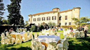 Una recepción de boda al aire libre con mesas redondas cubiertas de manteles blancos y sillas, instalada en una zona cubierta de césped con un edificio grande al fondo.
