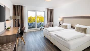 Ein Hotelzimmer mit Schreibtisch, zwei Betten, einem Sessel, einem Tisch und Blick auf Bäume durch das Fenster.