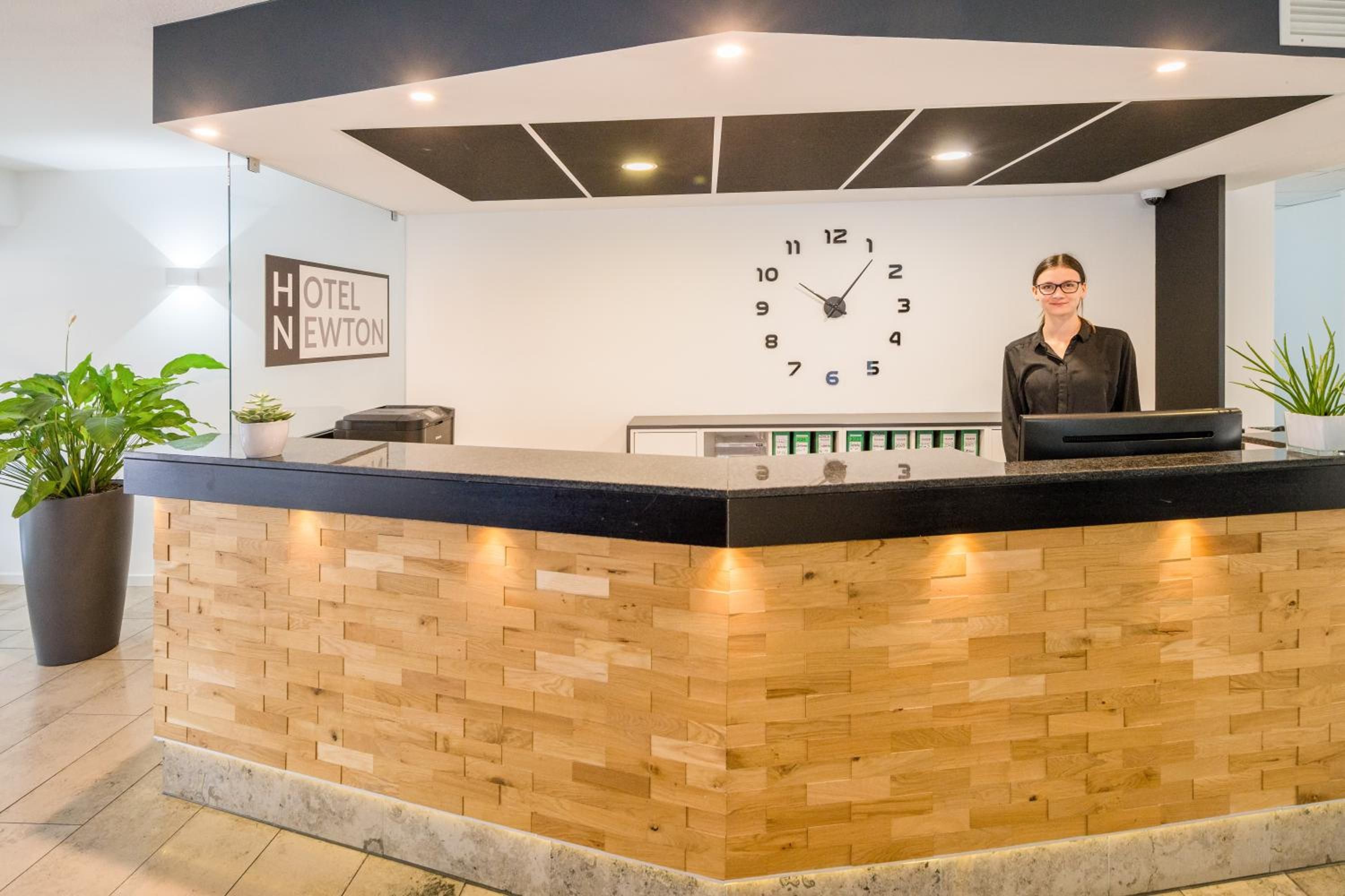 Ein Hotelrezeptionstresen mit einer Uhr, einem Schild mit der Aufschrift 'HOTEL NEWTON' und einer Rezeptionistin.