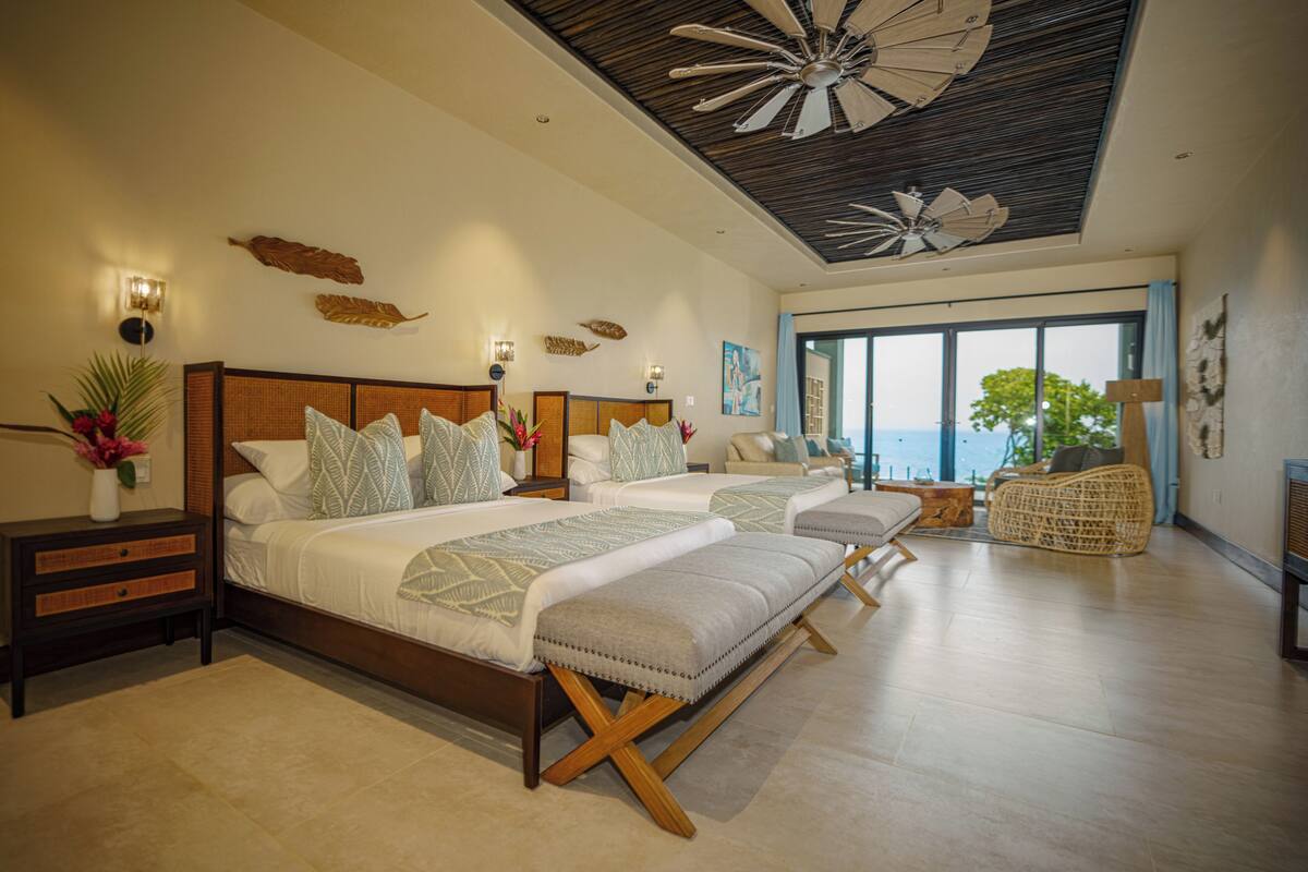 A spacious bedroom with two beds, a ceiling fan, and a large window offering a view of the sea.