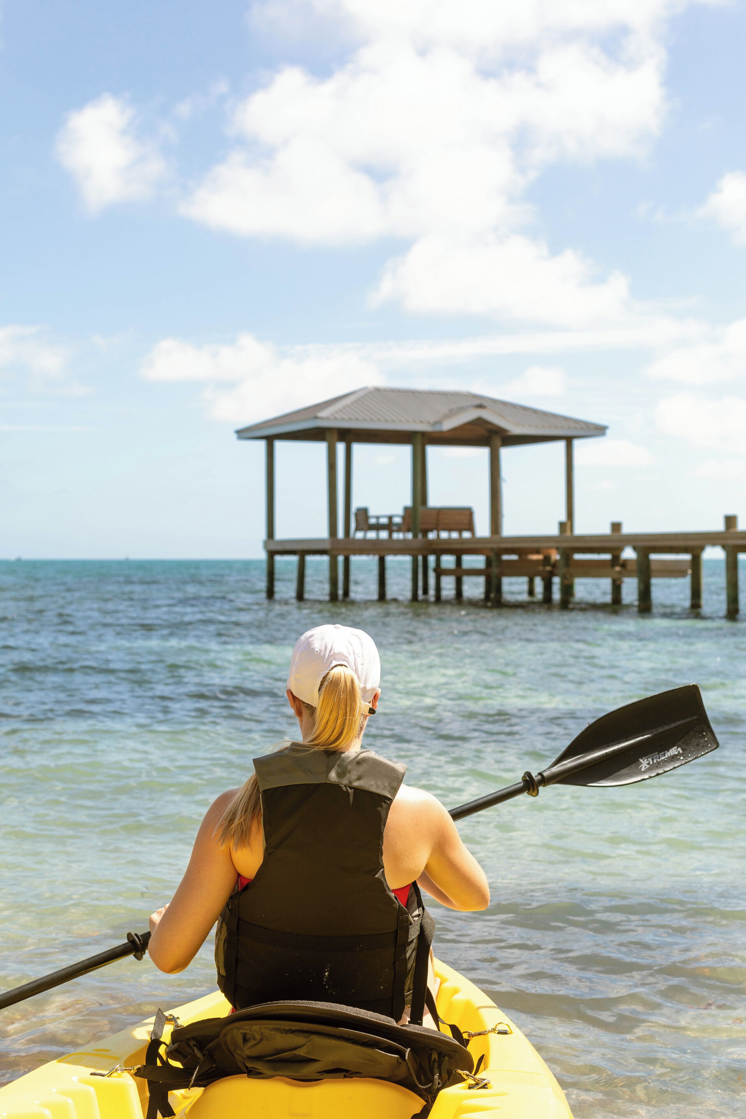 Una persona haciendo kayak en un día despejado, con un muelle de madera y un pabellón al fondo.